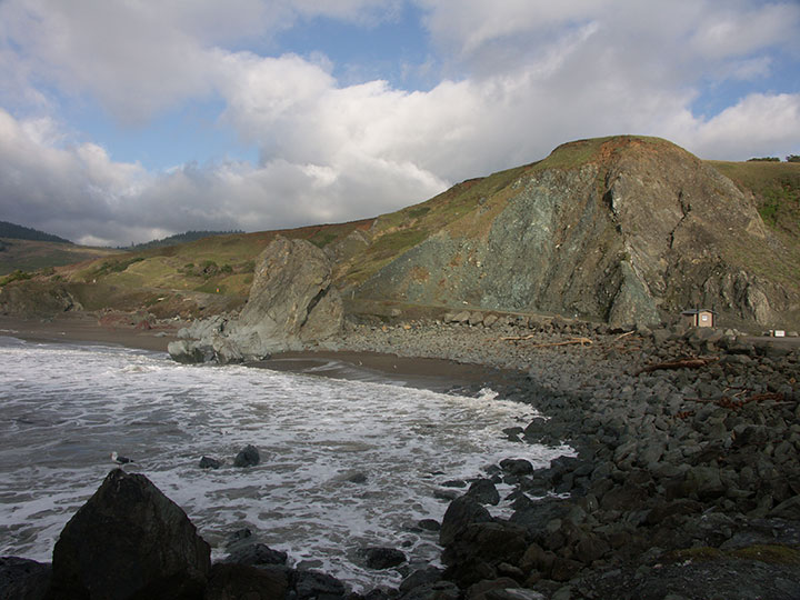 grey and red rocks are topped with green grass, the cliffsides meet a small road with some state beach signage and a restroom, the ocean breaks on large rocks creating a bed of sea foam along a dark sand beach
